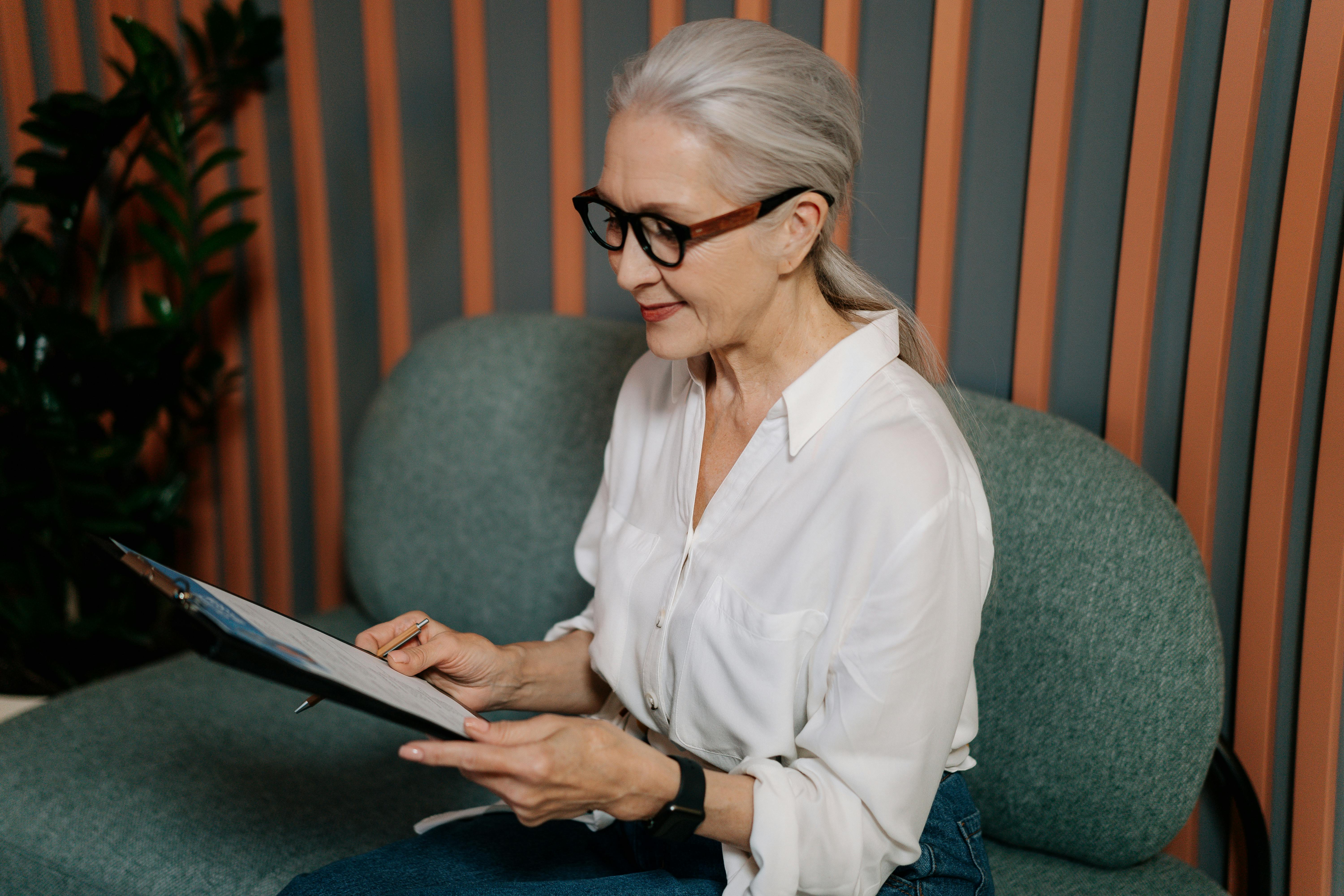 pexels-photo-5989942-5989942 Elderly woman in glasses reviewing documents on clipboard, sitting indoors with a focused expression.
